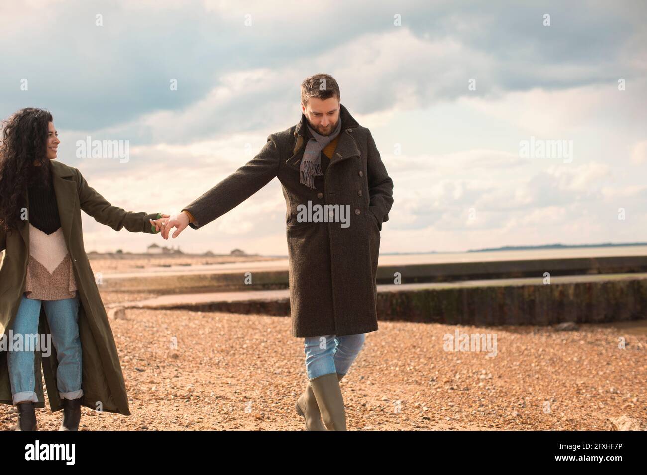 Paar in Wintermänteln, die Hände halten und am sonnigen Strand spazieren gehen Stockfoto