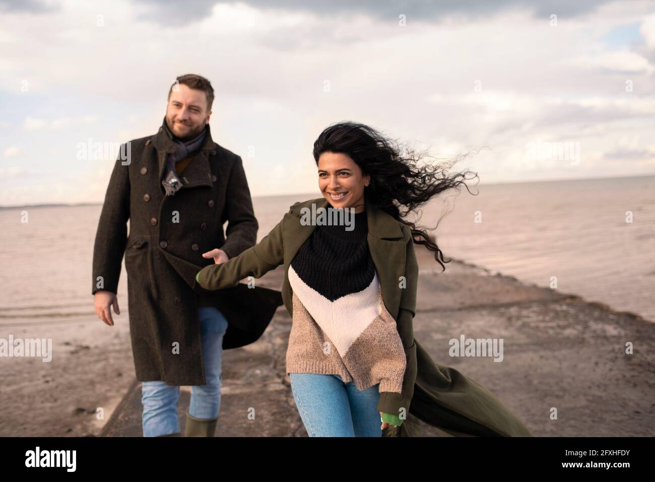 Glückliches Paar in Wintermänteln, das die Hände am Meeressteg hält Stockfoto