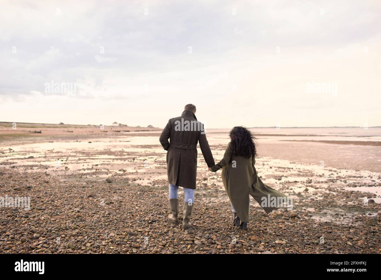 Paar in Wintermänteln, die sich die Hände halten und am Strand spazieren gehen Stockfoto
