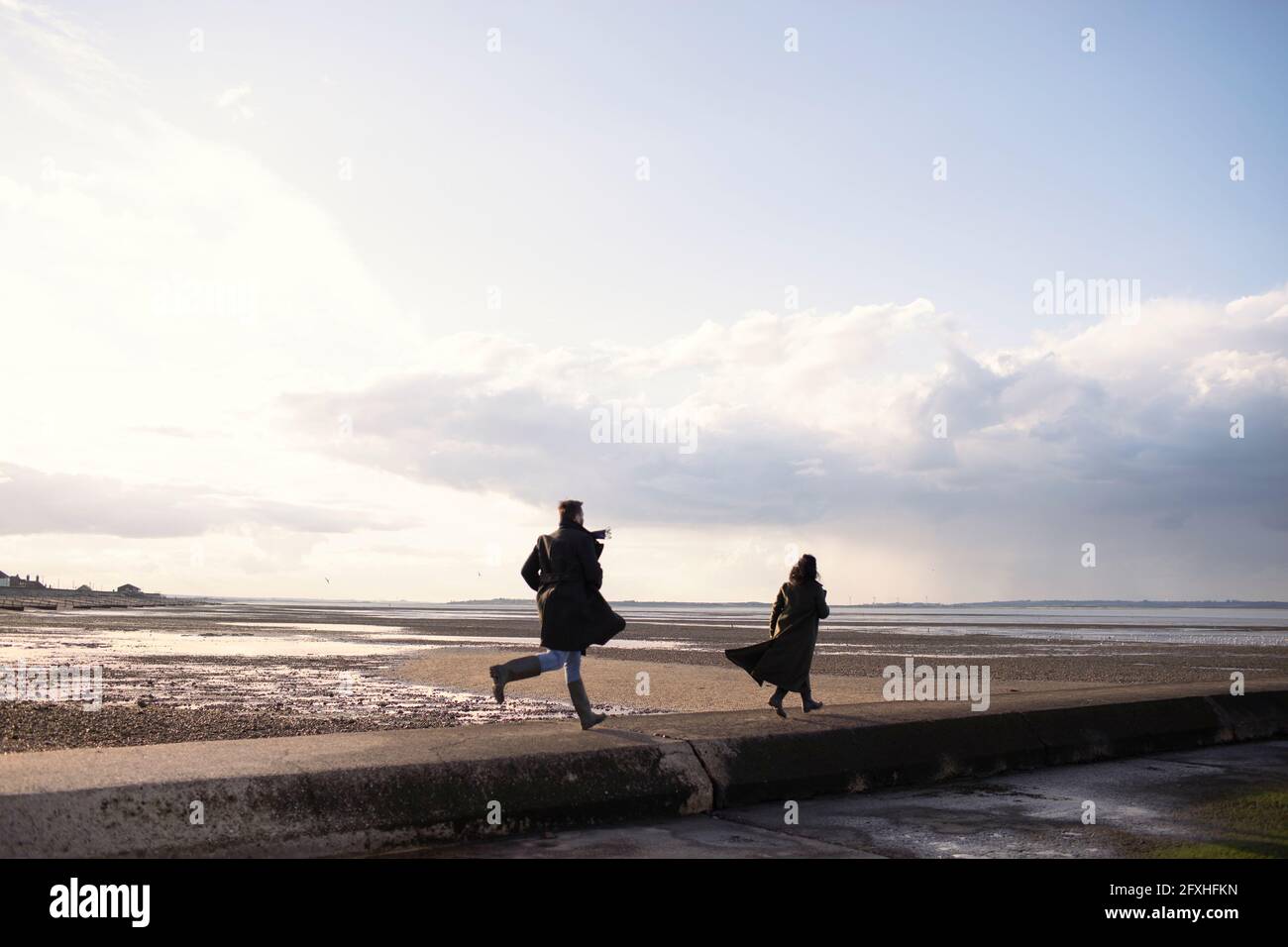 Paar in Wintermänteln, die auf einem sonnigen Anlegesteg am Strand laufen Stockfoto