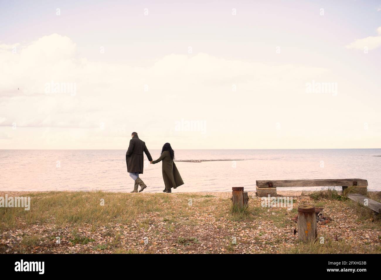 Paar in Wintermänteln, die sich die Hände halten und am Strand spazieren gehen Stockfoto
