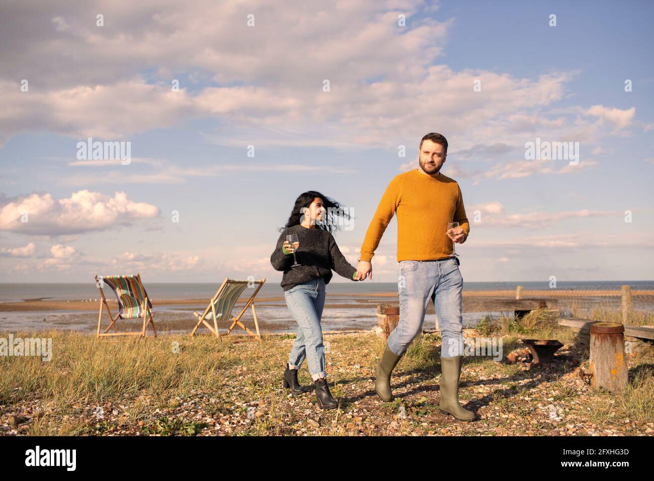 Ein glückliches Paar mit Wein, der die Hände hält und am sonnigen Strand spazieren geht Stockfoto