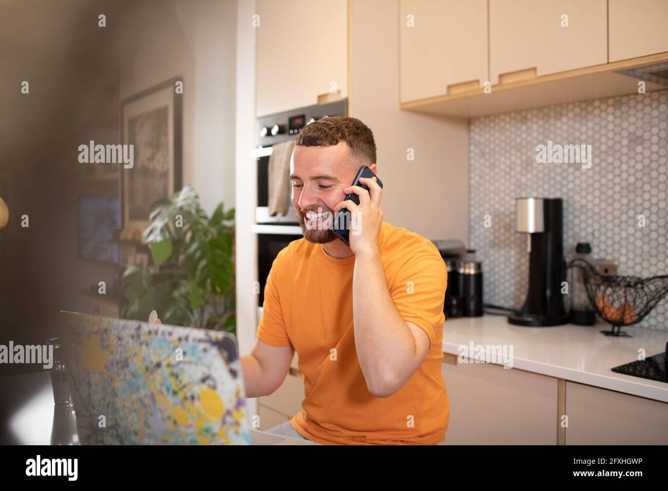 Glücklicher junger Mann, der von zu Hause aus mit einem Smartphone am Laptop spricht Stockfoto