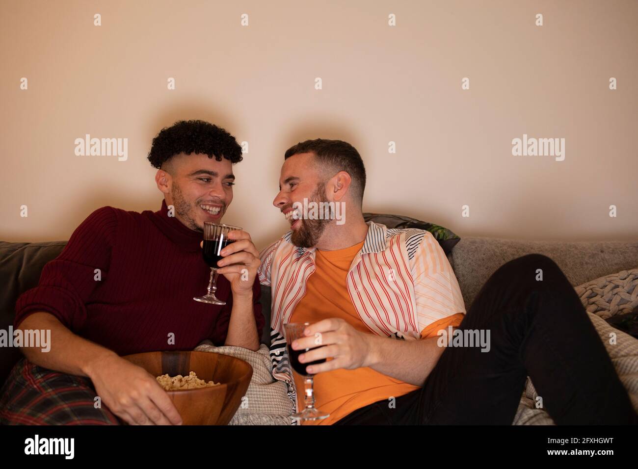 Glückliches schwules männliches Paar, das zu Hause Rotwein trinkt und Popcorn isst Stockfoto