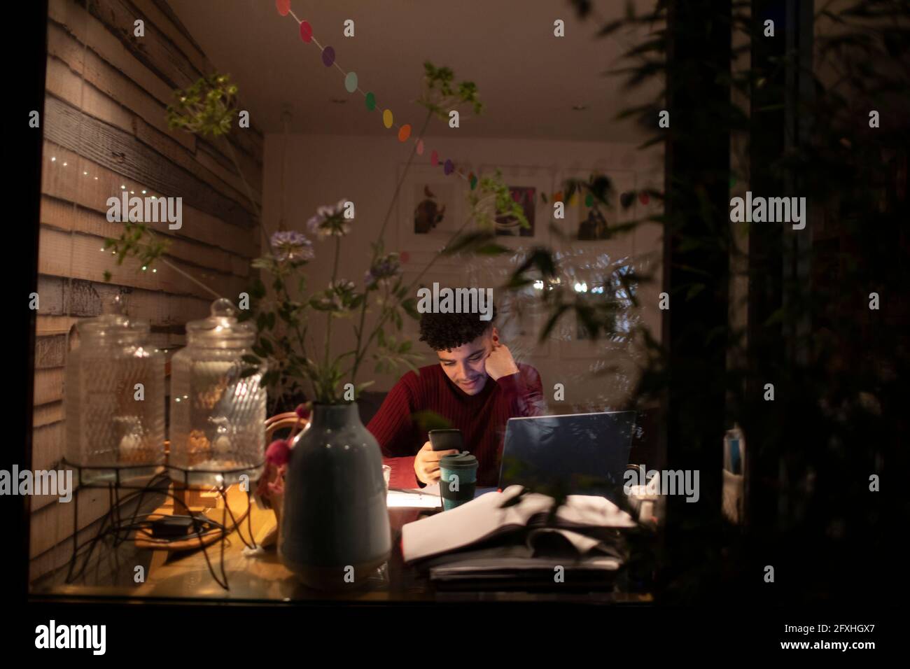 Junger Mann mit Smartphone, der nachts von zu Hause aus am Laptop arbeitet Stockfoto