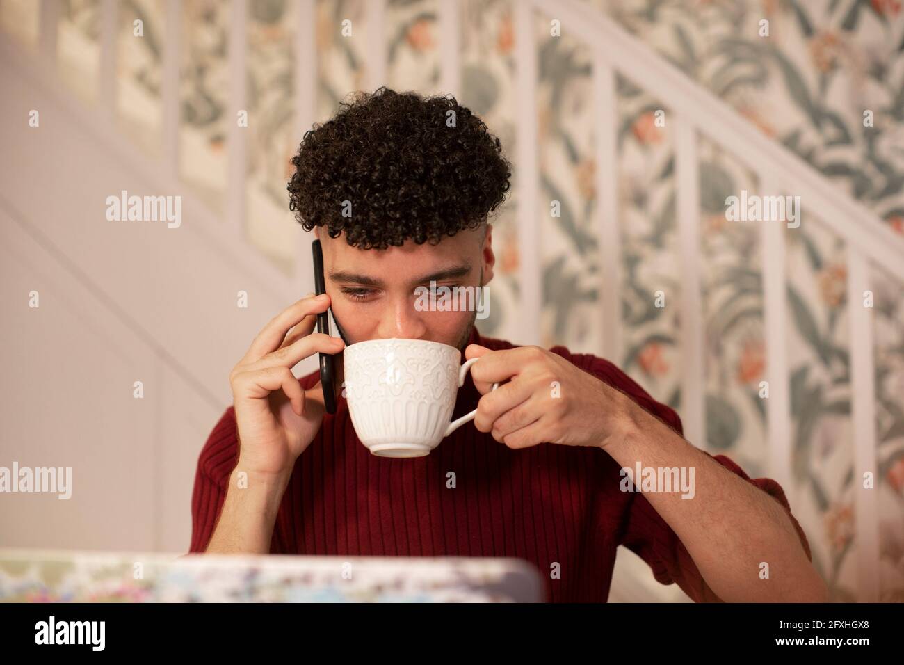 Junger Mann mit Kaffee, der am Laptop am Smartphone telefoniert Stockfoto