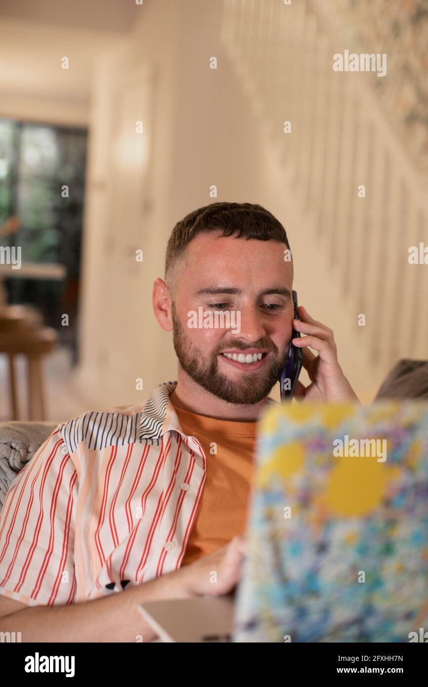Lächelnder junger Mann, der am Laptop mit dem Smartphone spricht Stockfoto