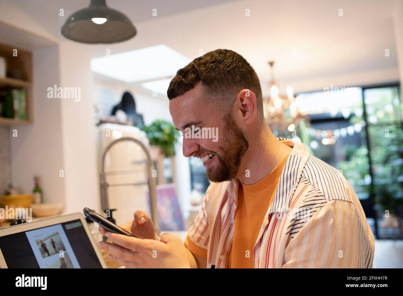 Glücklicher junger Mann, der Smartphone am Laptop in der Küche benutzt Stockfoto