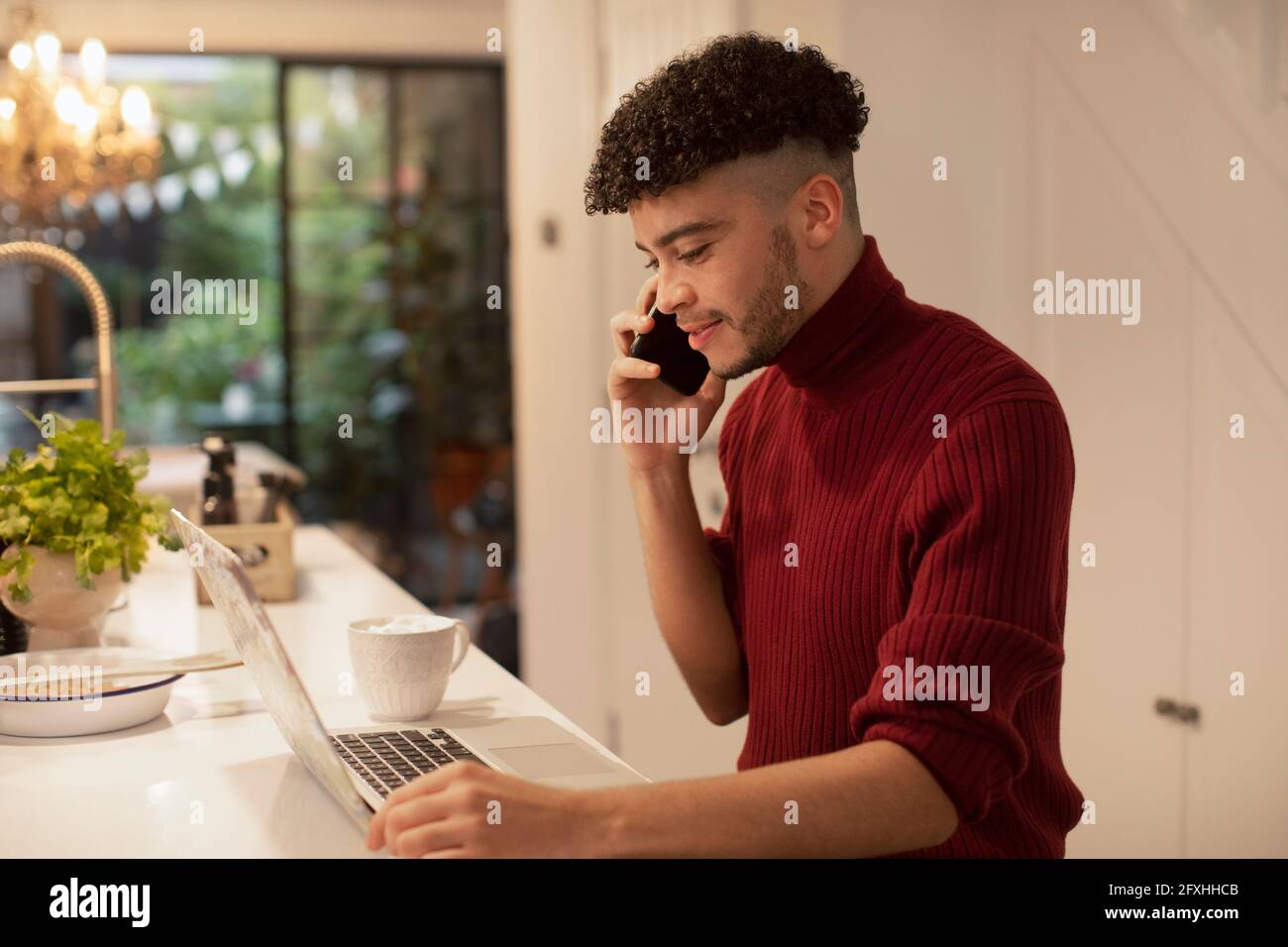 Junger Mann, der von zu Hause aus am Laptop mit einem Smartphone spricht Stockfoto