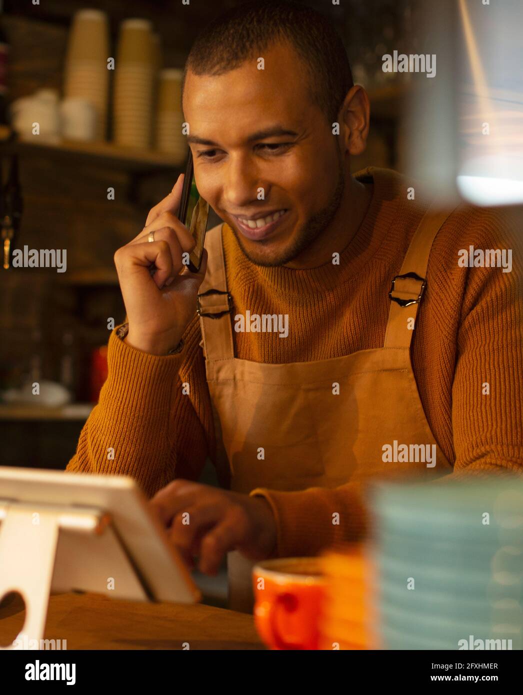 Lächelnder, männlicher Barista, der am digitalen Tablet im Café telefoniert Stockfoto