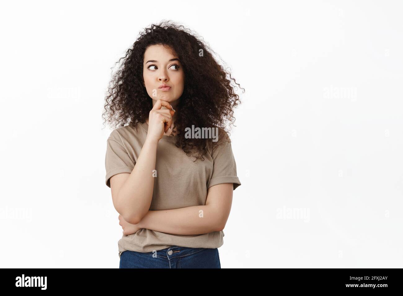 Nachdenkliche junge, moderne Frau berührt das Kinn, denkt über etwas nach, denkt nach und schaut beiseite, steht vor weißem Hintergrund Stockfoto