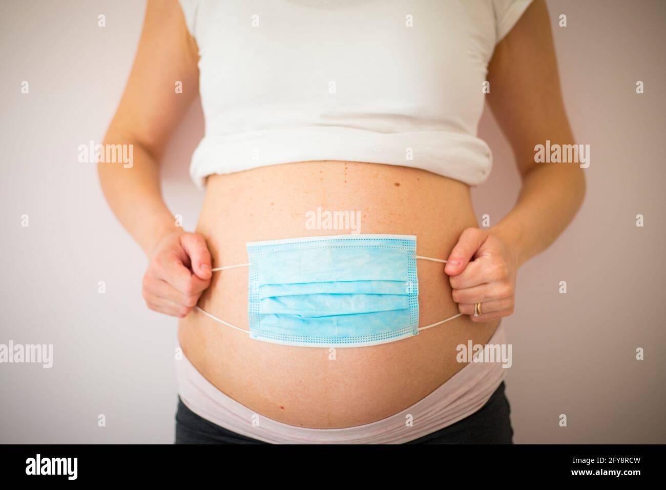 Während der Covid-19-Pandemie legte eine Schwangere eine blaue Gesichtsmaske auf den Bauch. Coronavirus in der Schwangerschaft Problem, Schutz Symbol. Vorderansicht, Zentralansicht. Stockfoto