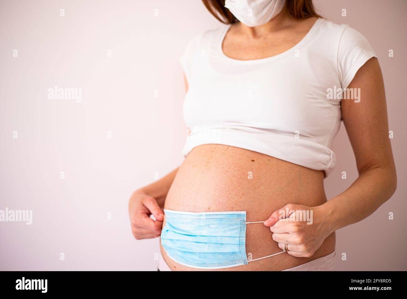 Eine Schwangere, die eine Gesichtsmaske trägt und während der Pandemie von Covid-19 eine weiße Gesichtsmaske auf den Bauch legt. Coronavirus in der Schwangerschaft Problem, Schutz Symbol. Stockfoto