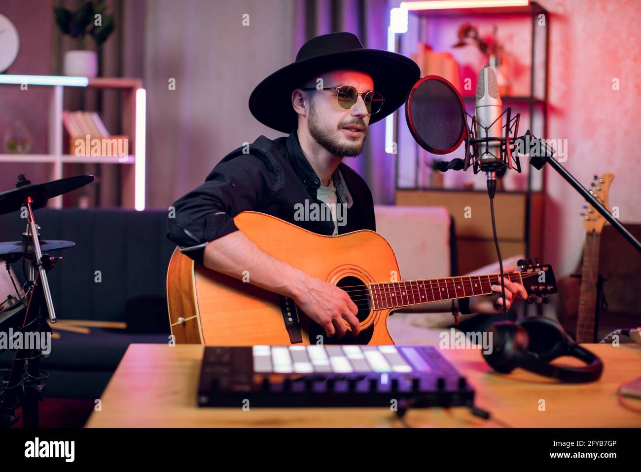 Professioneller männlicher Künstler mit Elektrogitarre und Mikrofon für die Aufnahme neuer Songs im Studio. Schöner Mann mit stilvollem schwarzen Hut und Brille. Stockfoto