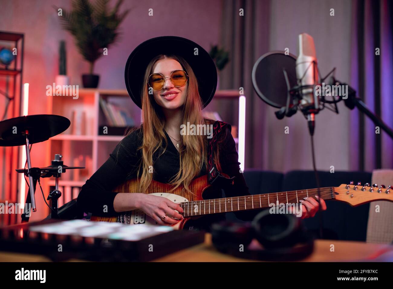 Lächelnde junge Frau mit Hut und Brille bei einer Probe im modernen Studio. Hübsche Künstlerin, die Gitarre spielt und im Mikrofon singt. Erstellung eines neuen Songs. Stockfoto