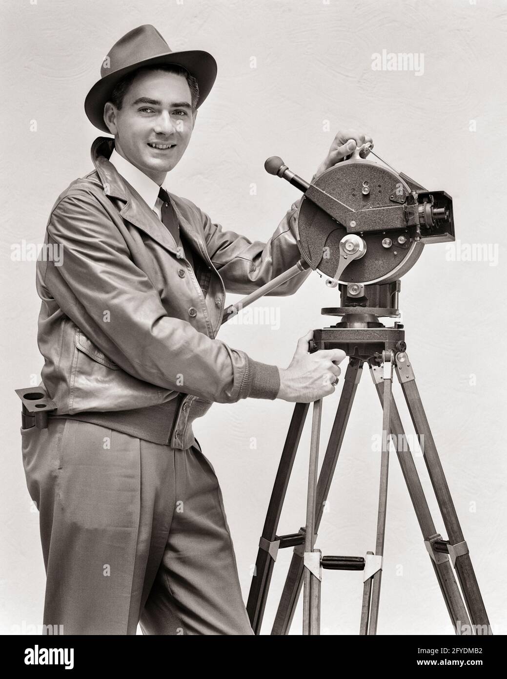 LÄCHELNDER MANN AUS DEN 1930ER JAHREN, KAMERAMANN, DER DIE HANDKURBEL-FILMKAMERA AKELEY BEDIENTE BLICK AUF DIE KAMERA TRÄGT LEDERJACKE FEDORA HUT - U153 HAR001 HARS KARRIERE TECHNOLOGIE INFORMATIONEN FREUDE LIFESTYLE JOBS STUDIO SHOT MOTION DREHARBEITEN KOPIEREN RAUM HALBE LÄNGE PERSONEN INSPIRATION MÄNNER KAMERAMANN BERUF VERTRAUEN TRAGBARE STATIV B & W AUGENKONTAKT KURBEL FEDORA GESCHICKLICHKEIT BERUF GLÜCK FÄHIGKEITEN BETRIEB FRÖHLICH ABENTEUER STRATEGIE KARRIERE WAHL WISSEN RICHTUNG ARBEITERSTOLZ AUF AUTORITÄT BESCHÄFTIGUNG BERUFE LÄCHELT VERBINDUNG KONZEPTUELL IMAGINATION FREUDVOLLE STILVOLLE MITARBEITERJACKE AUS LEDER Stockfoto