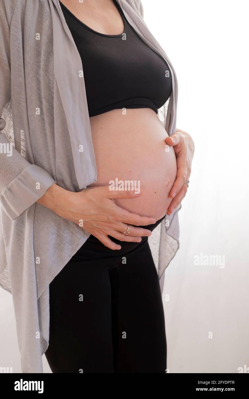 Eine Schwangere, die in Schwarz gekleidet ist, mit einer langen grauen Jacke, wobei ihr Bauch mit den Händen auf ihrem Bauch vor einem weißen, vertikalen Hintergrund freigelegt ist Stockfoto