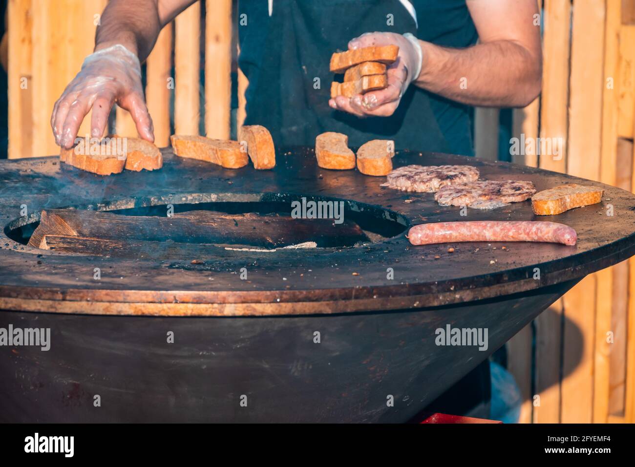 Die Hände eines Mannes mit Handschuhen, der Würstchen, Steaks und Brotscheiben auf einem großen Grill im Freien röstet. Grillfest im Stadtpark. Stockfoto