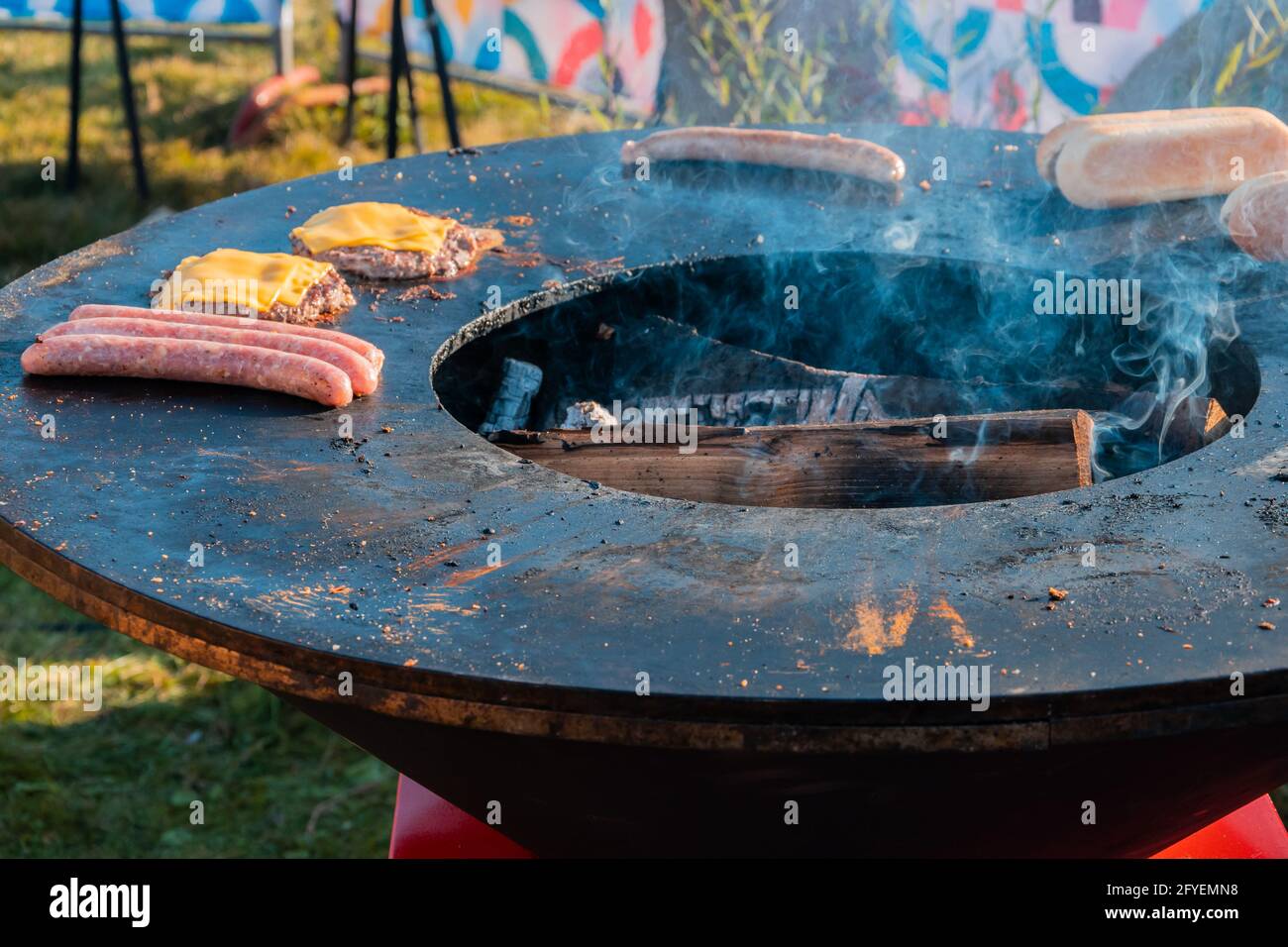 Auf dem großen runden Holzgrill im Freien werden Würstchen, Hotdog-Brötchen, Rinderpasteten mit Käse für Burger gebraten. Grillfest in der Stadt p Stockfoto