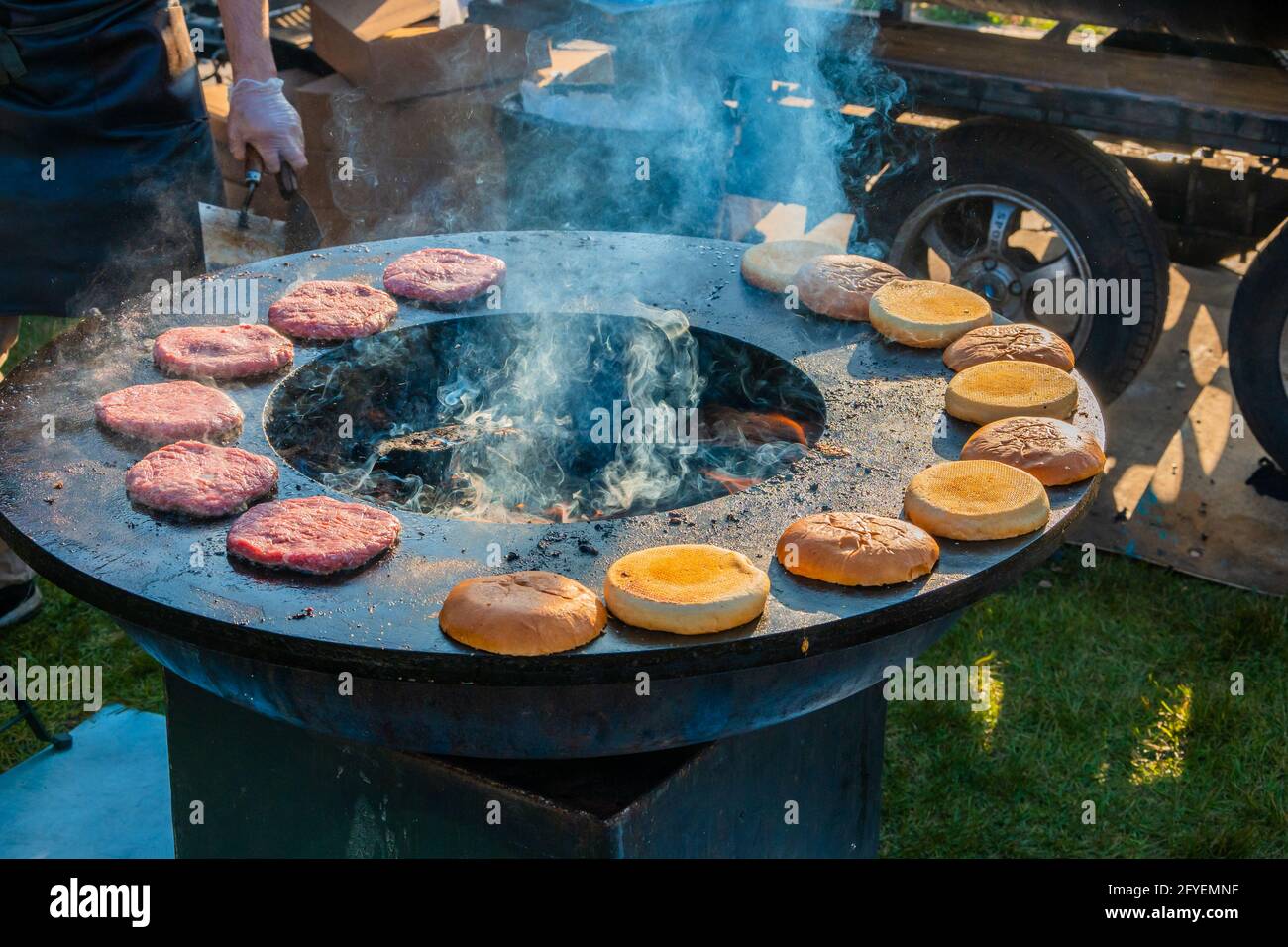 Auf einem großen, runden Holzgrill draußen werden Rindersteaks und Burger-Brötchen gegrillt. Grillfest im Stadtpark. Street Fast Food. Stockfoto