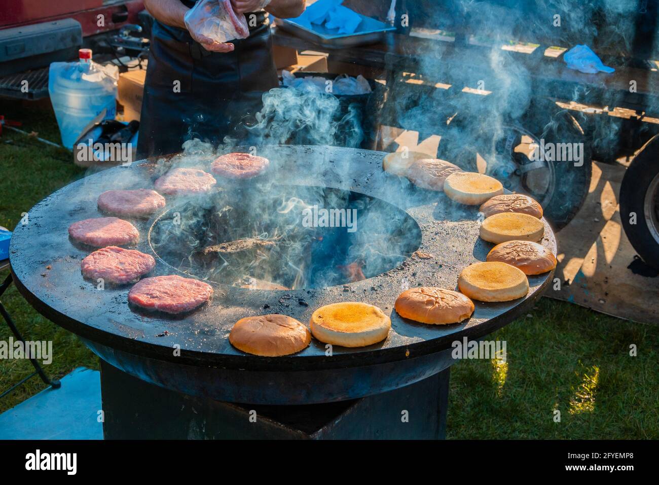 Auf einem großen, runden Holzgrill draußen werden Rindersteaks und Burger-Brötchen gegrillt. Grillfest im Stadtpark. Street Fast Food. Stockfoto