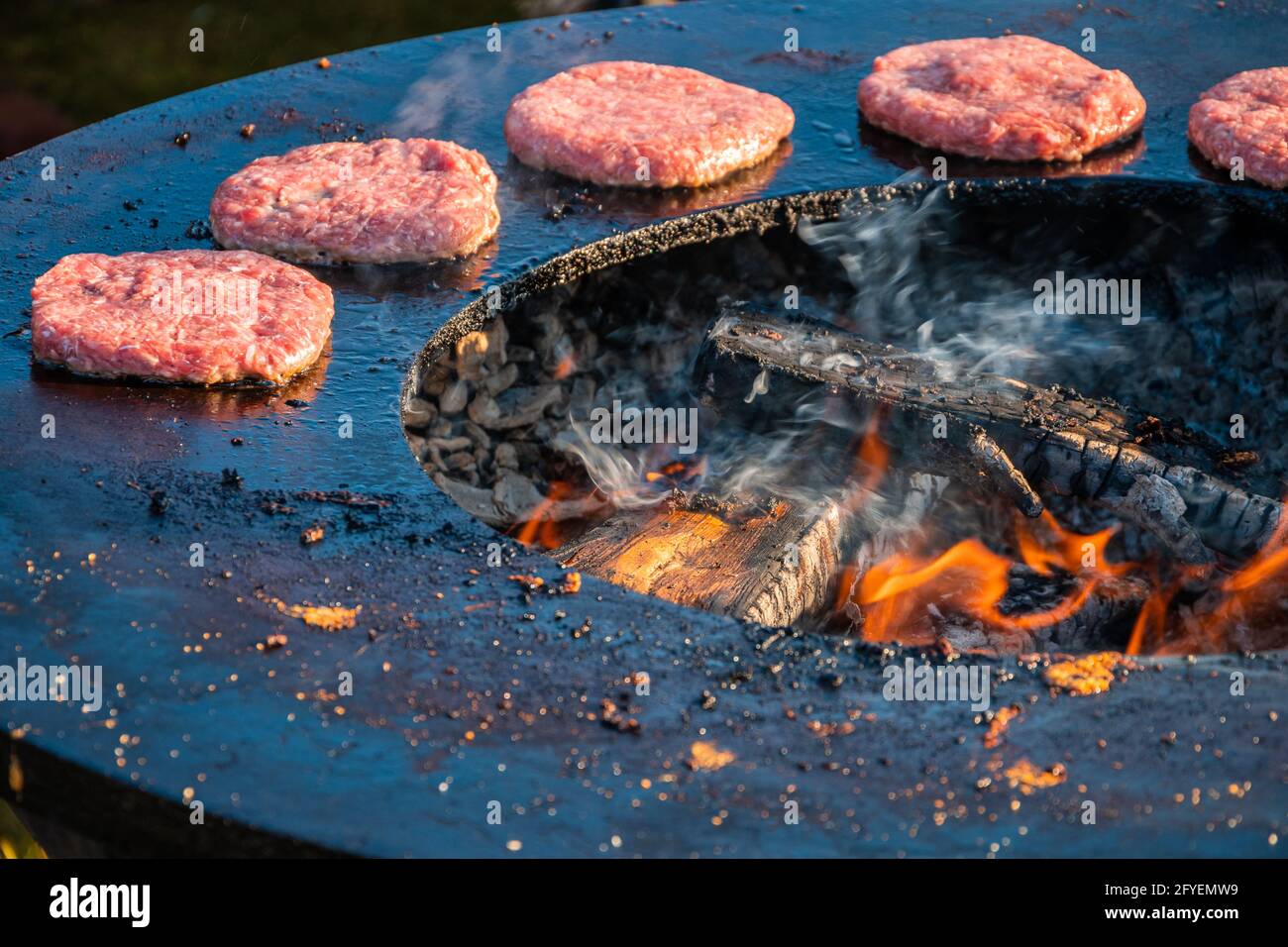 Auf einem großen runden Holzgrill werden Rindersteaks für Burger gegrillt. Nahaufnahme. Grillfest im Stadtpark. Street Fast Food. Stockfoto