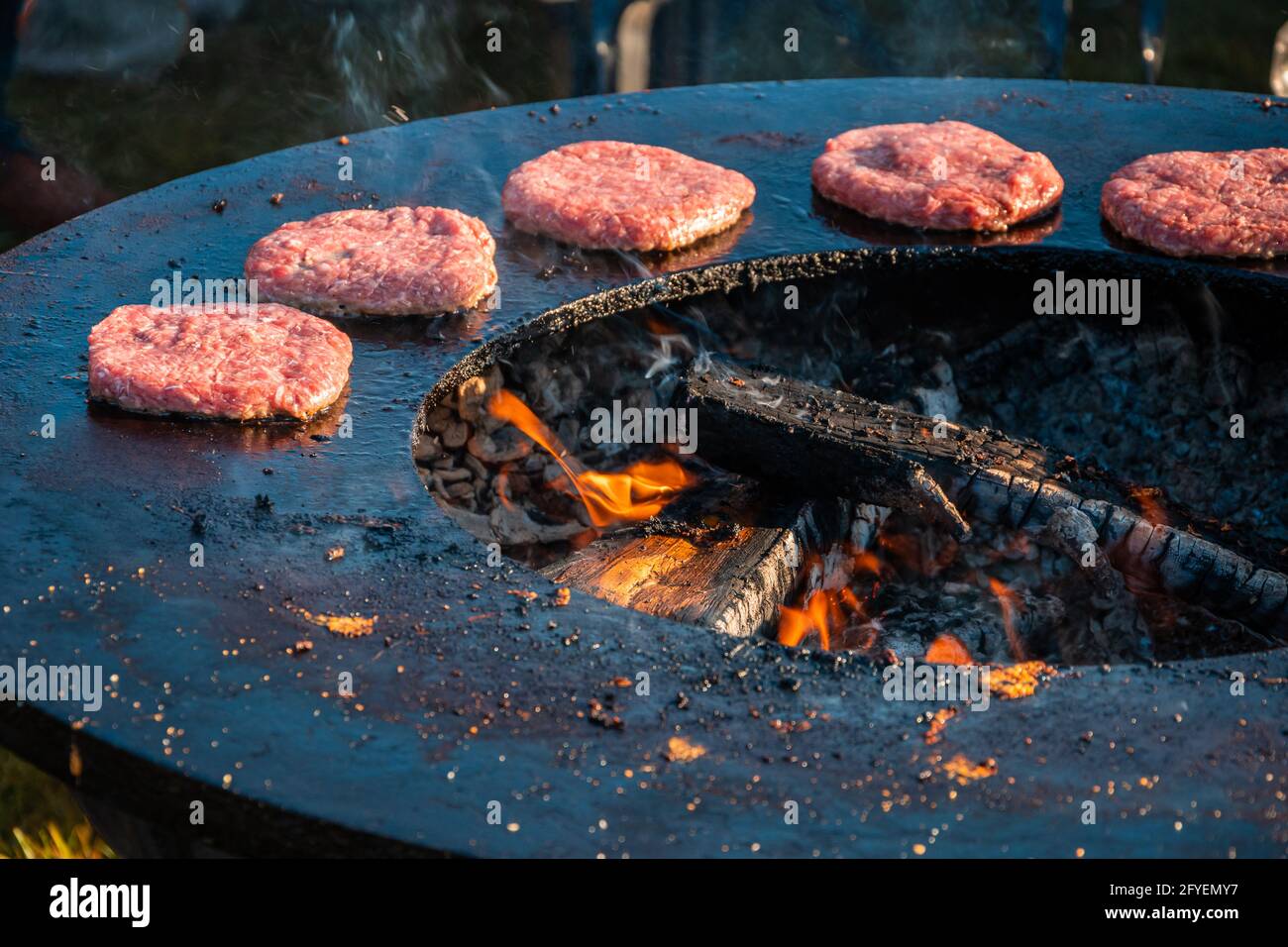 Auf einem großen runden Holzgrill werden Rindersteaks für Burger gegrillt. Nahaufnahme. Grillfest im Stadtpark. Street Fast Food. Stockfoto