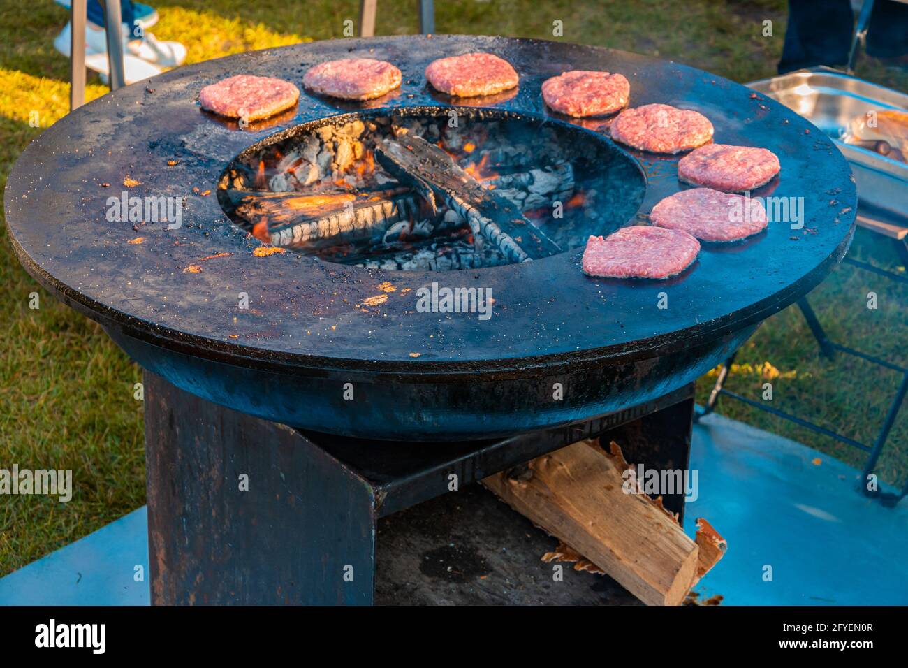 Auf dem Rasen röstet ein großer runder Holzgrill Rindersteaks für Burger. Grillfest im Stadtpark. Street Fast Food. Stockfoto
