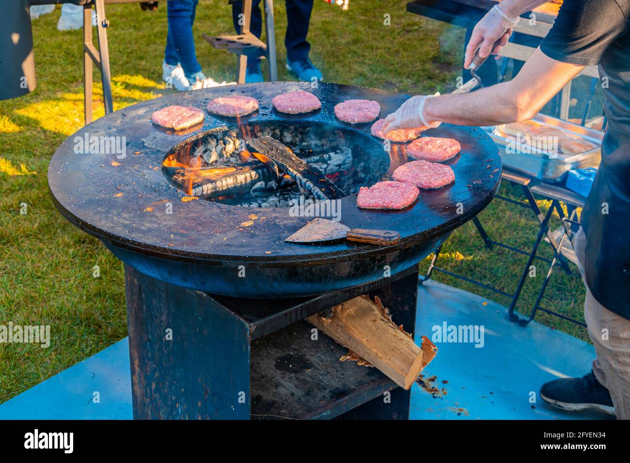 Auf dem Rasen röstet ein großer runder Holzgrill Rindersteaks für Burger. Grillfest im Stadtpark. Street Fast Food. Stockfoto