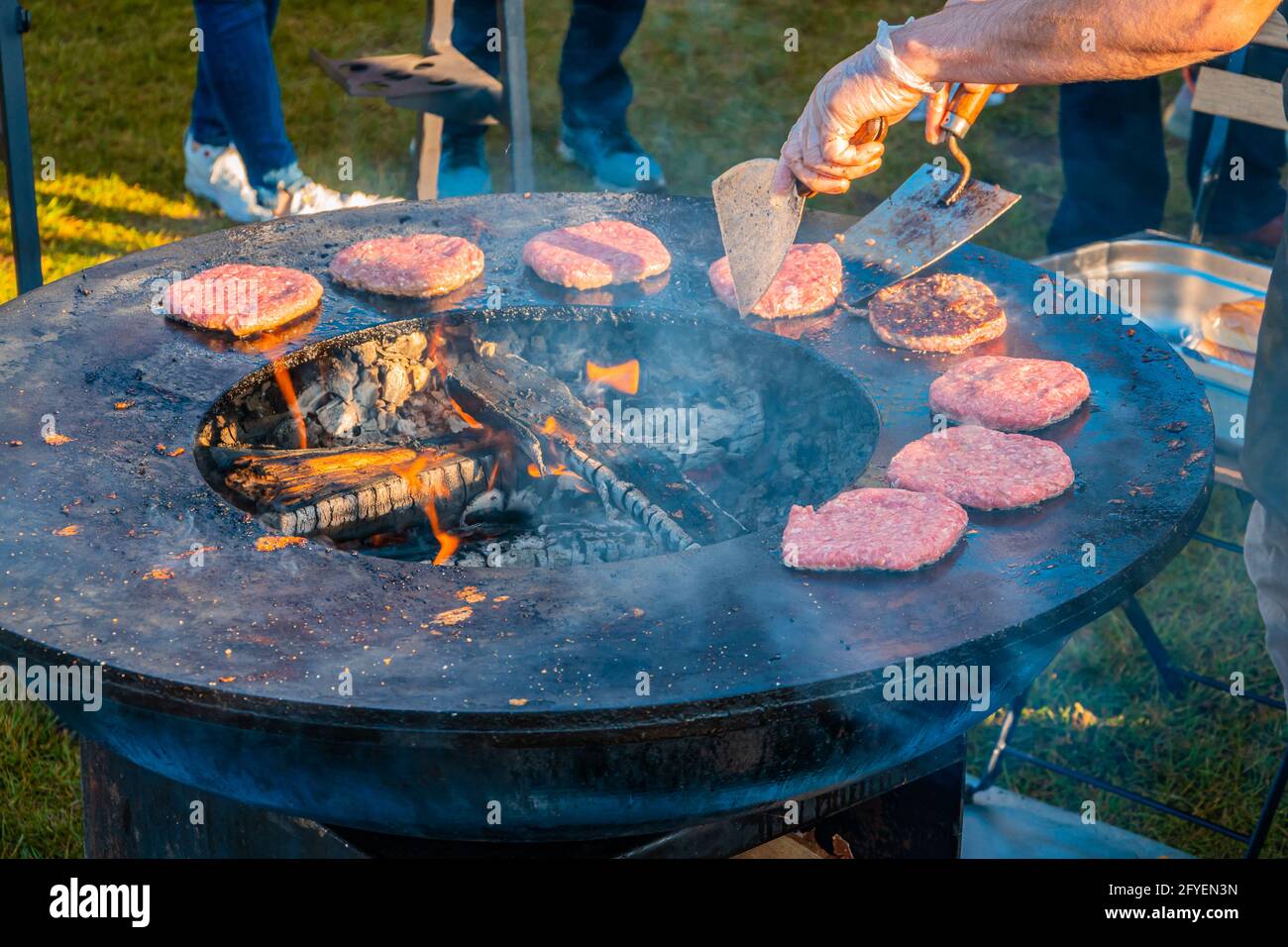 Auf dem Rasen röstet ein großer runder Holzgrill Rindersteaks für Burger. Der Koch dreht die gebratenen Koteletts um. Grillfest in der CI Stockfoto