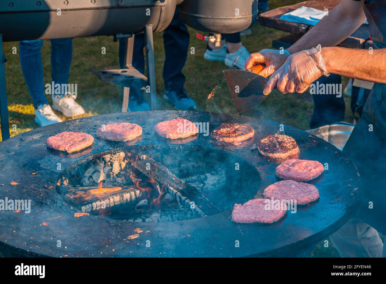 Auf dem Rasen röstet ein großer runder Holzgrill Rindersteaks für Burger. Grillfest im Stadtpark. Street Fast Food. Stockfoto