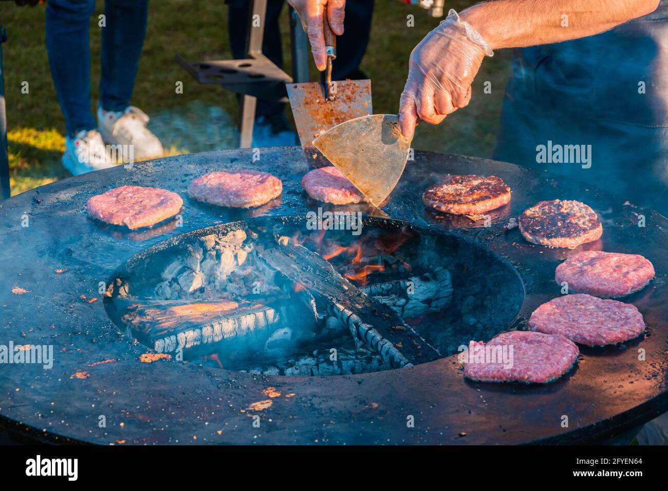 Auf dem Rasen röstet ein großer runder Holzgrill Rindersteaks für Burger. Der Koch dreht die gebratenen Koteletts um. Grillfest in der CI Stockfoto