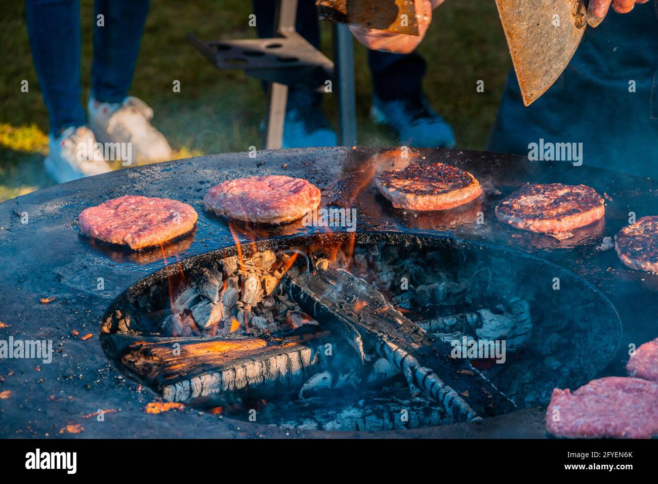 Auf dem Rasen röstet ein großer runder Holzgrill Rindersteaks für Burger. Grillfest im Stadtpark. Street Fast Food. Stockfoto