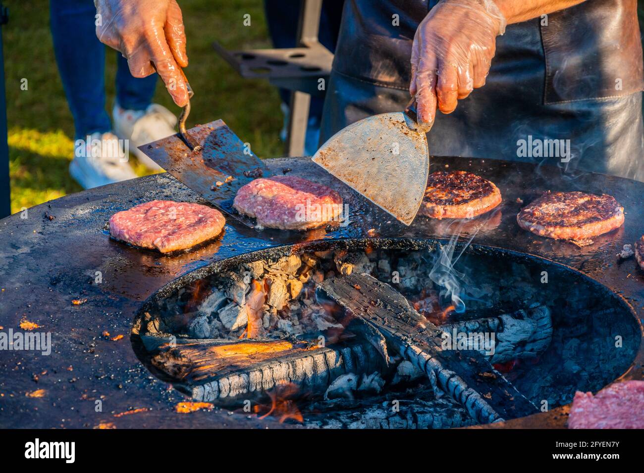 Auf dem Rasen röstet ein großer runder Holzgrill Rindersteaks für Burger. Der Koch dreht die gebratenen Koteletts um. Grillfest in der CI Stockfoto