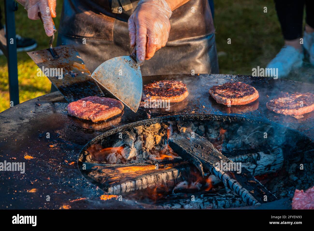 Auf dem Rasen röstet ein großer runder Holzgrill Rindersteaks für Burger. Ein Koch in einer Lederschürze dreht die frittierten Schnitzel um. Grill f Stockfoto