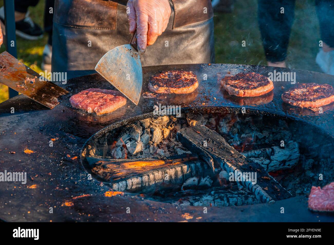 Auf dem Rasen röstet ein großer runder Holzgrill Rindersteaks für Burger. Ein Koch in einer Lederschürze dreht die frittierten Schnitzel um. Grill f Stockfoto