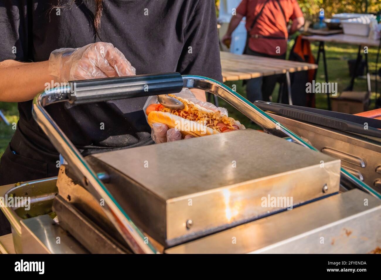 Die Handschuhen eines Küchenchefs, der einen frisch gerösteten Hot Dog mit Füllung füllt. Grillfest im Stadtpark. Street Fast Food. Stockfoto