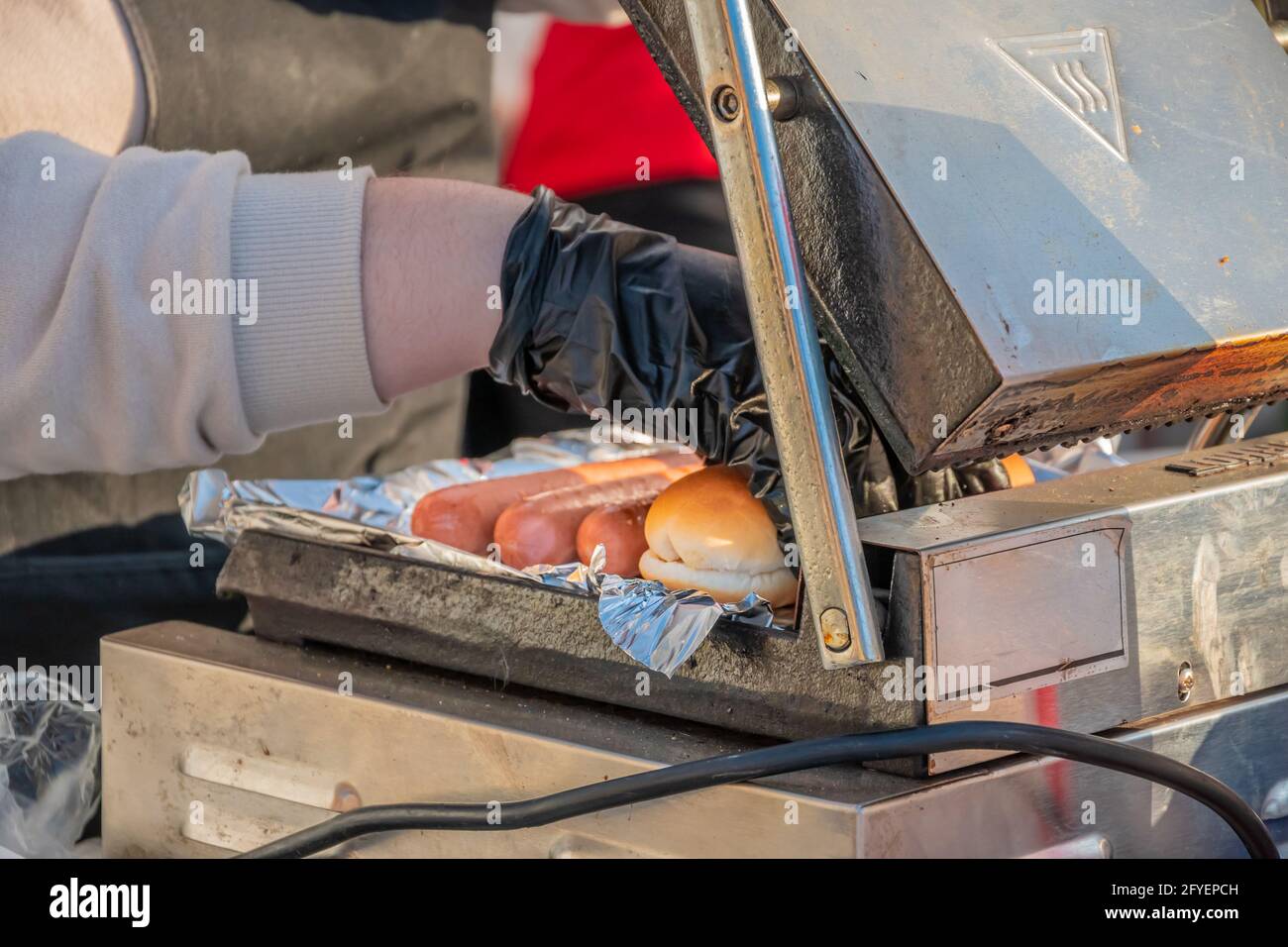 Der Koch nimmt eine auf einem rotierenden Schweißgerät gebratene Hot-Dog-Wurst. Grillfest im Stadtpark. Street Fast Food. Stockfoto