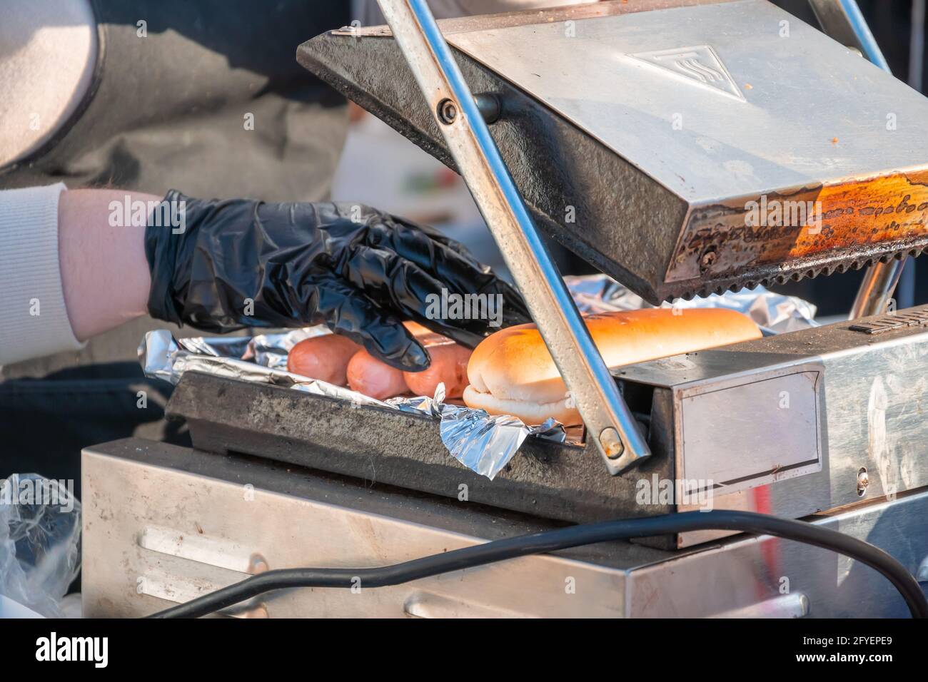 Der Koch nimmt eine auf einem rotierenden Schweißgerät gebratene Hot-Dog-Wurst. Grillfest im Stadtpark. Street Fast Food. Stockfoto