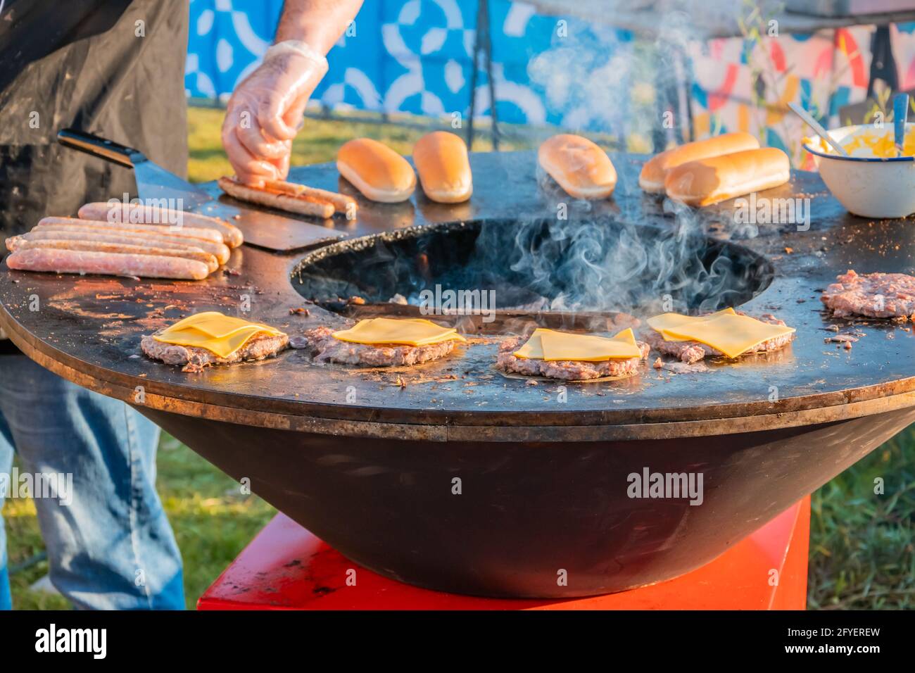 Auf dem Rasen röstet ein großer runder Holzgrill Rindersteaks für Burger. Der Koch dreht die gebratenen Koteletts um. Grillfest in der CI Stockfoto