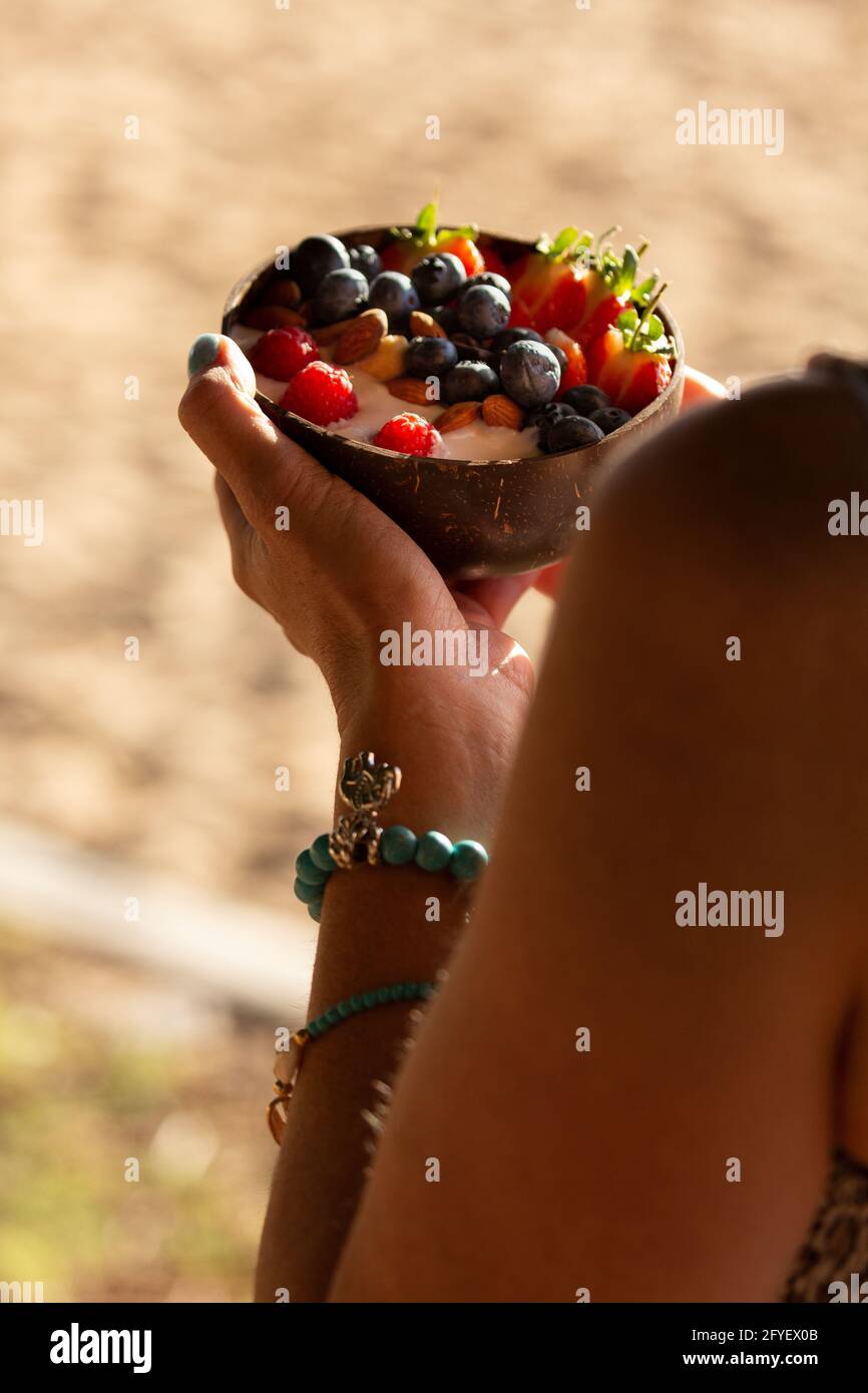 Eine Frau hält morgens in einer Kokosnussschale am Strand eine köstliche, geglättete Frühstückschüssel mit frischen Sommerfrüchten, Joghurt und Müsli. Stockfoto