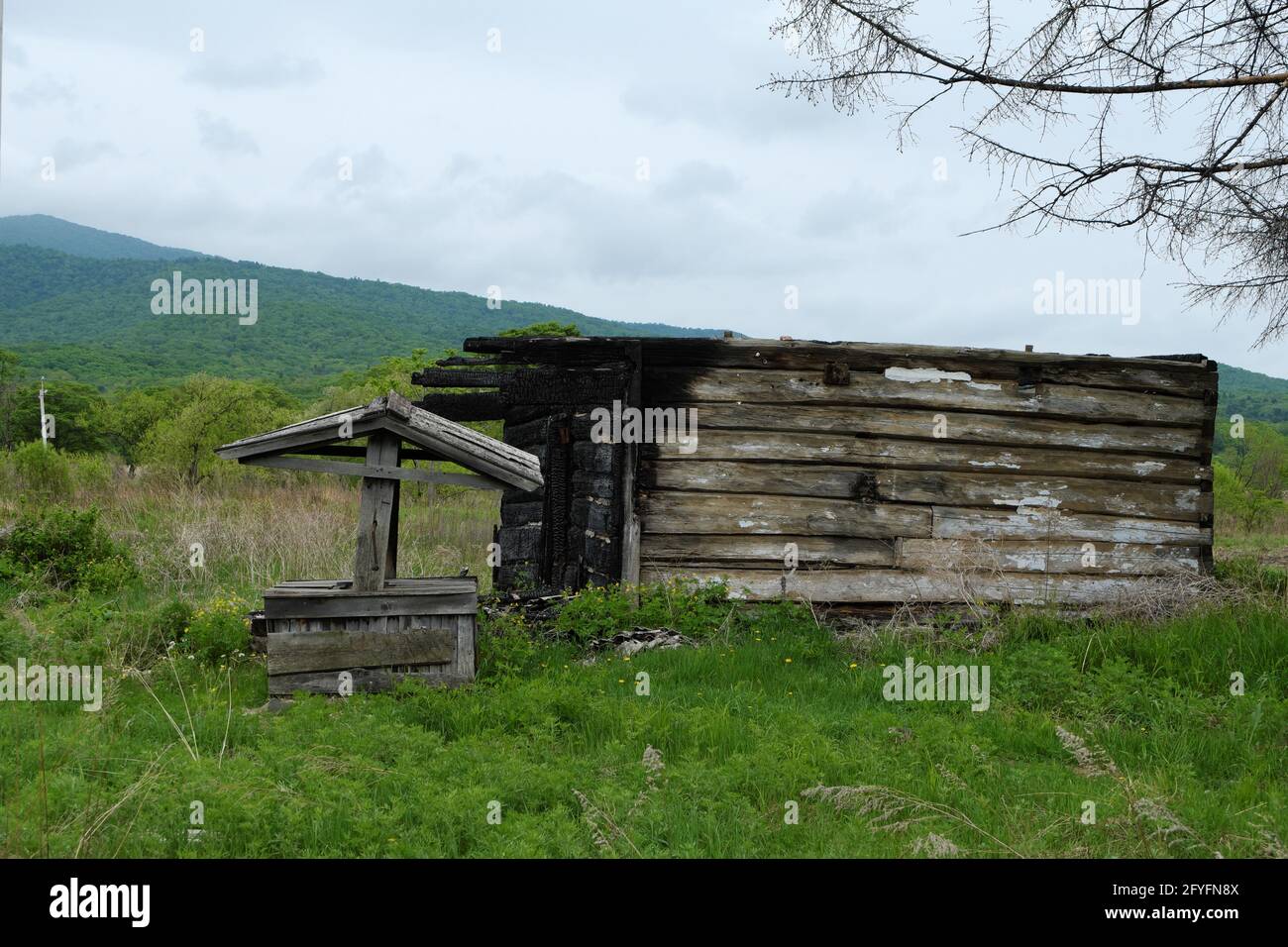 Die Überreste eines verlassenen ausgebrannten ländlichen Hauses mit einem Brunnen in einer Lichtung in einer bergigen Gegend. Stockfoto