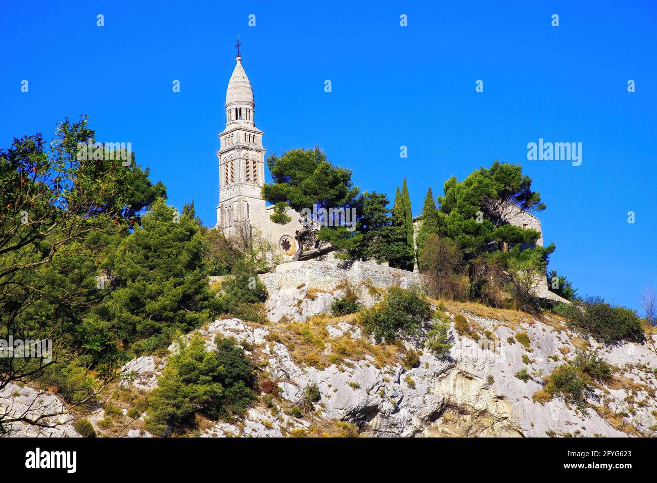 Tiefwinkelansicht auf isolierter römisch-katholischer Kirche auf Berggipfel mit Kiefern- und Zypressbäumen gegen blauen Himmel - Plan d'Orgon, Bouche du Rhône, Nachgewiesen Stockfoto