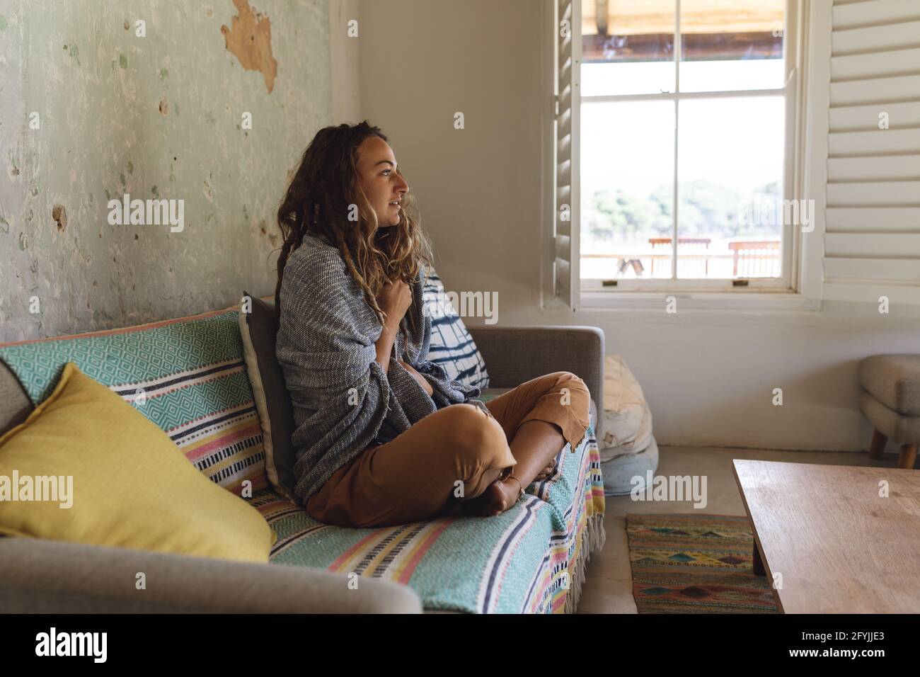 Glückliche kaukasische Frau, die eine Decke über den Schultern auf dem Sofa trägt Im sonnigen Cottage Wohnzimmer Stockfoto