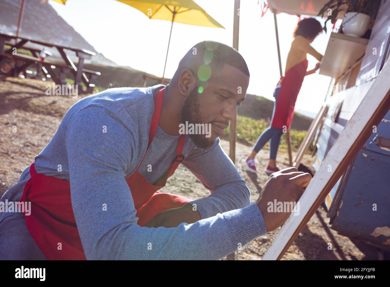 Lächelnder afroamerikanischer Mann schreibt auf der Menütafel daneben Foodtruck an sonnigen Tagen Stockfoto