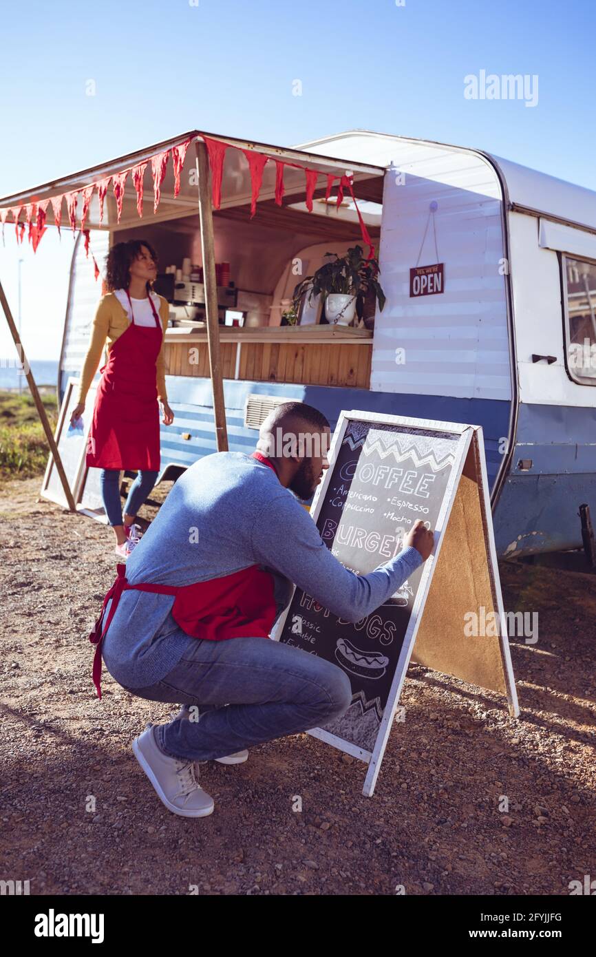 Vielfältiges Paar, das am sonnigen Tag einen Food Truck am Meer öffnet und vorbereitet, Mann, der auf der Speisekarte schreibt Stockfoto
