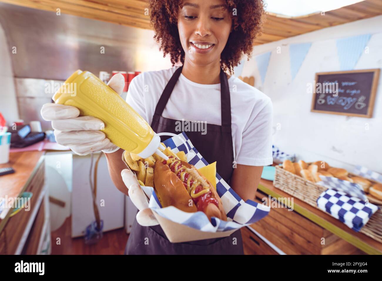 Lächelnde Mischling Frau Senf auf Hot Dog hinter Zähler im Foodtruck Stockfoto