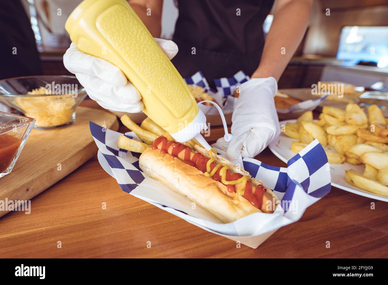 Mittelteil einer Frau mit gemischter Rasse, die Senf auf einen Hot Dog gießt Hinter dem Tresen im Foodtruck Stockfoto