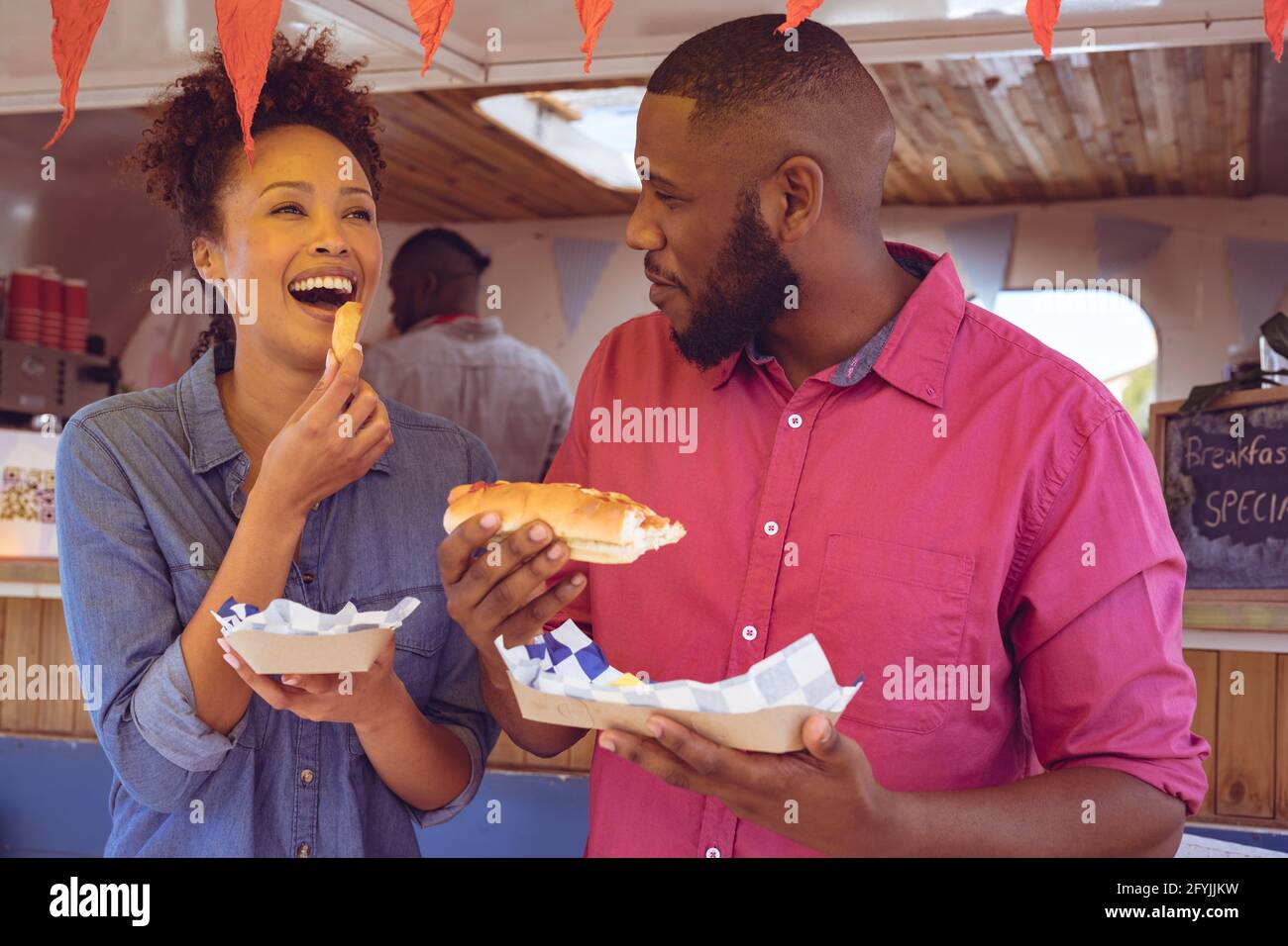 Lächelndes, vielfältiges Paar, das Hot Dog und Kartoffelkeile isst Foodtruck an sonnigen Tagen Stockfoto