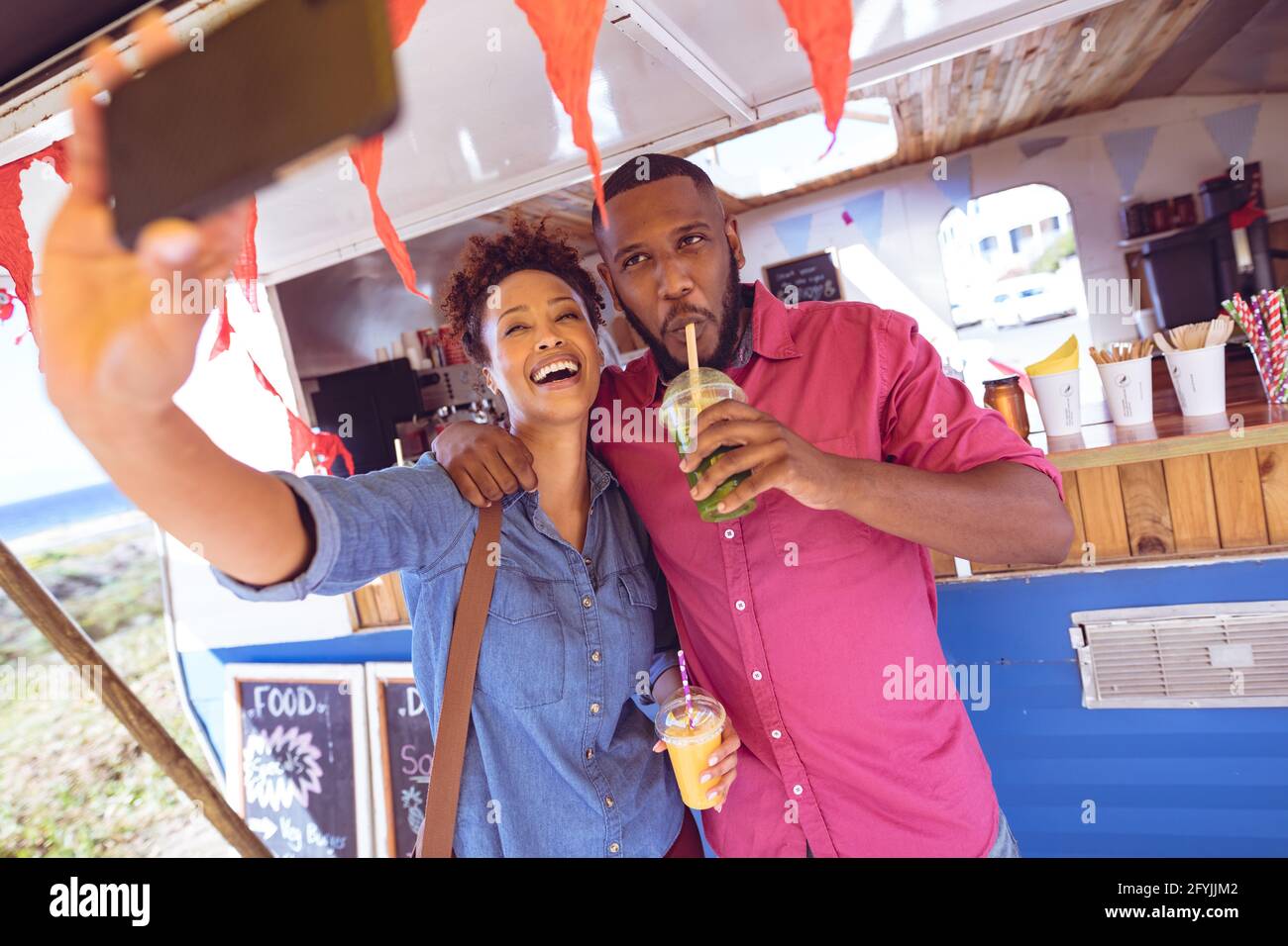 Lächelndes, vielfältiges Paar, das Selfie mit dem Smartphone gemacht hat und etwas getrunken hat Mit dem Foodtruck am Meer Stockfoto